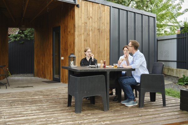 A family gather around a black table on a wooden deck of tiny house, sharing drinks and laughter in a stylish backyard. The surrounding greenery complements the contemporary design of the space