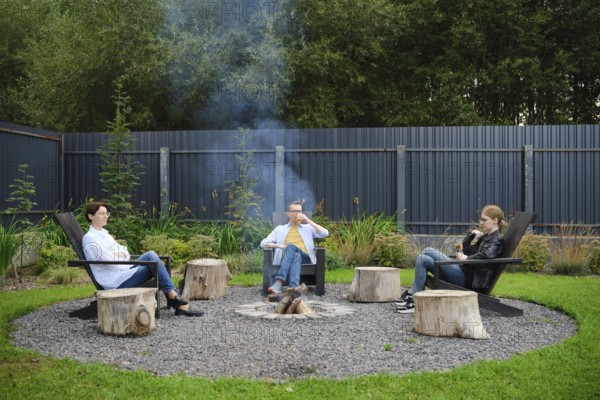 A family relax around a circular fire pit, seated on rustic chairs during a weekend. Smoke drifts gently in the air as they enjoy each other's company in a peaceful garden filled with greenery