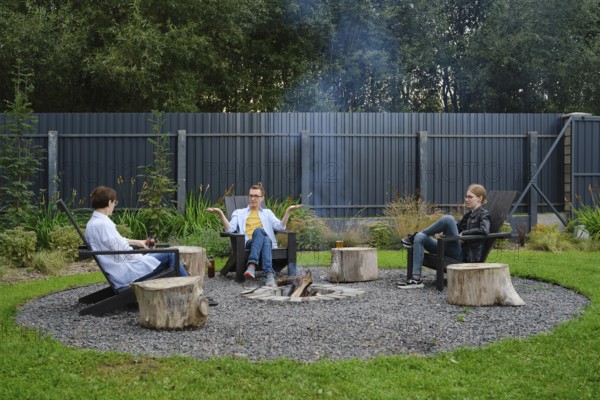 A family enjoy a relaxing evening around a fire pit in a backyard. They are sitting on wooden stools, engaged in conversation while the gentle smoke rises into the cool evening air