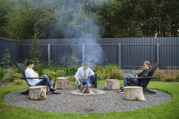 A family sits comfortably on wooden chairs, gathering around a fire pit in a spacious garden. Smoke rises from the fire as they engage in warm conversation, surrounded by greenery
