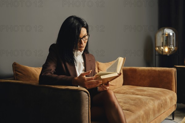 A person with glasses is deeply engrossed in reading a book while seated on a stylish couch in a contemporary living room. The ambient lighting casts a warm glow, creating a cozy atmosphere