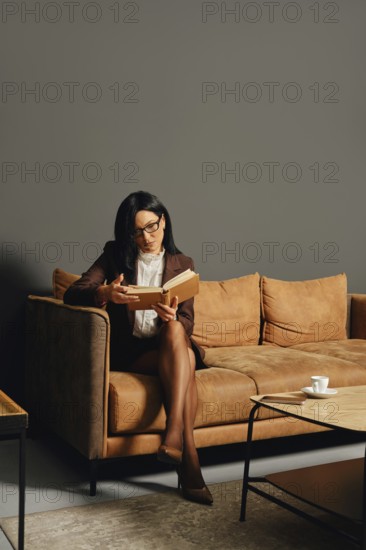 A businesswoman sits on a stylish brown couch in an office and reading a book during the break