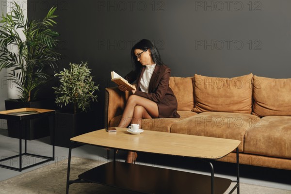 A woman dressed in a smart outfit is enjoying a quiet moment with a book on a cozy couch. The modern living room features greenery and minimalist furniture, creating a calm atmosphere