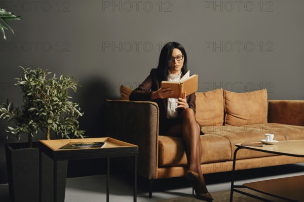 A woman in professional attire sits comfortably on a brown sofa, reading a book intently. A small table nearby holds a coffee cup and a potted plant, enhancing the cozy atmosphere in the room