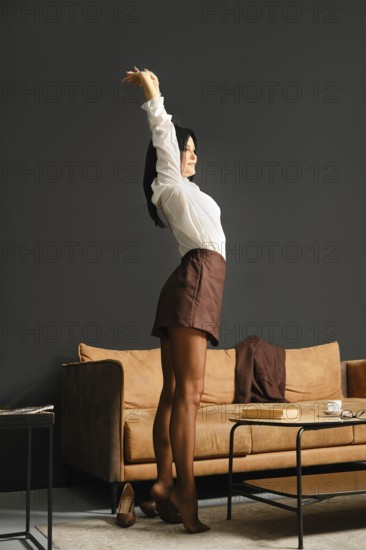 A slender woman dressed in a white blouse and a brown skirt stands on tiptoe and stretching arms in a stylish living room