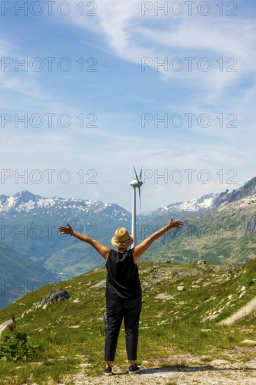 Woman with Arms Outstretched Standing in Front of a Wind Turbine with Mountain in the Swiss Alps and Glacier and Against Blue Sky and Clouds in a Sunny Summer Day in Andermatt, Uri, Switzerland