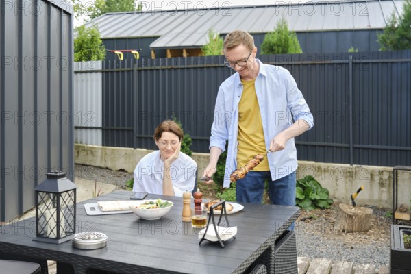 Middle-aged couple enjoy a barbecue meal outdoors in a backyard. Man is serving skewers on a grill, while his wife watches happily. The setting is sunny, with plants and outdoor furniture around
