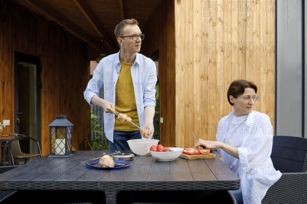 During a sunny day a couple is engaged in preparing a meal outdoors. One person is skewering food while the other slices tomatoes, both are distracted from their work and look away