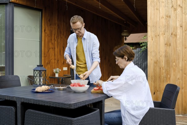 Two people enjoy preparing a meal in a vibrant outdoor space. One person is slicing vegetables while the other arranges food on a plate, surrounded by natural wood and modern decor