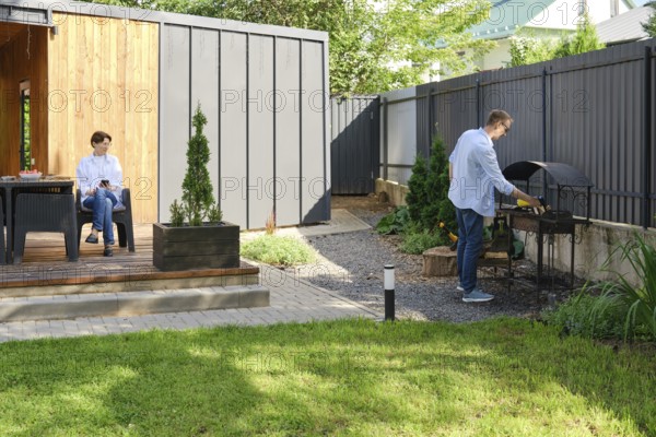 A couple spends the day outdoors at their contemporary home. The wife is seated on a patio, while her husband is grilling in the backyard, surrounded by greenery and wooden elements
