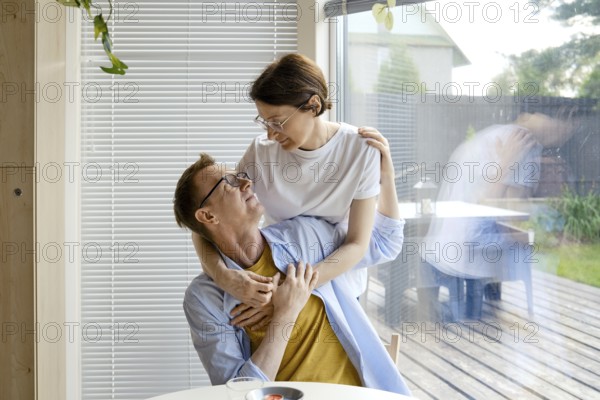 A man and woman share an affectionate moment indoors, surrounded by natural light. They smile at each other while the woman playfully embraces the man. A serene garden is visible outside the window