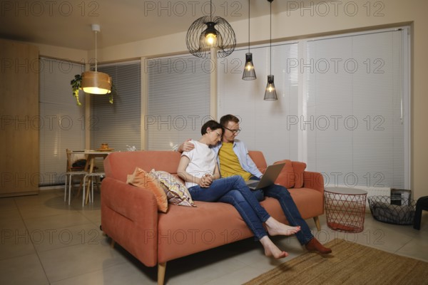 A husband embraces his wife while sitting on a couch in a modern living room. They are watching a new episode of the series online