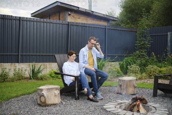 Two people enjoy a tranquil moment by the fire pit in a lush backyard. They share stories and meditatively watching the fire