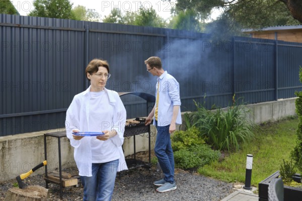 Two people are preparing a barbecue outdoors. A woman holds a plate while walking away, and a man cooks on the grill, surrounded by greenery and a fence