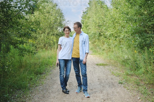 A middle-aged couple enjoys a stroll along a gravel path shaded by lush greenery. They smile at each other and have a leisurely conversation