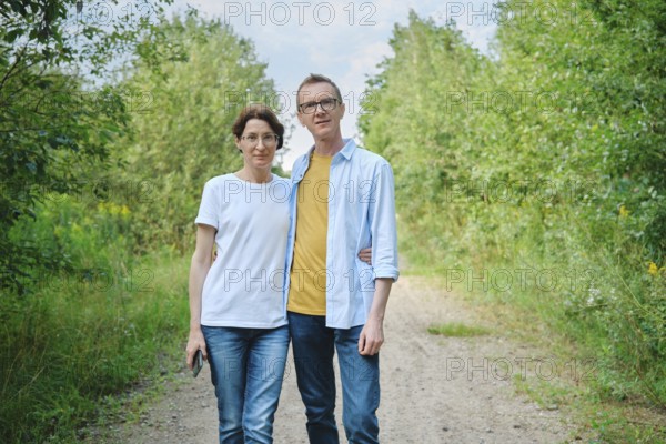 A middle-aged couple stands closely together on a natural path surrounded by lush greenery. They are casually dressed, enjoying a relaxed day outdoors with bright sunlight filtering through the trees