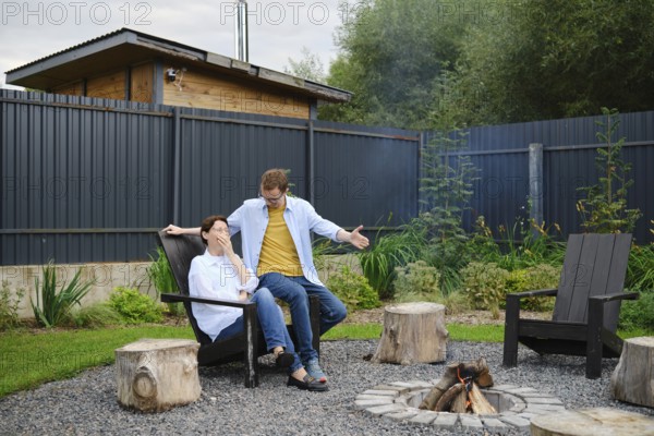 A couple spends a relaxed evening outdoors near a fire pit in their backyard. They share playful banter, surrounded by lush greenery and wooden seating, creating a warm and inviting atmosphere