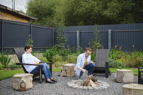 Middle-aged couple enjoy a relaxing evening in a backyard, sitting by a campfire surrounded by greenery. A husband kneels beside the logs and starts fire, while his wife sits comfortably in a chair