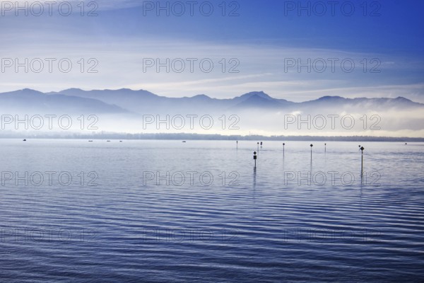 A calm Lake Constance with mountains and fog in the morning light under a blue sky, Lindau