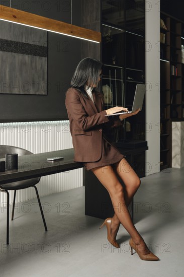A businesswoman in a brown suit is sitting on a desk while focused on her laptop in a contemporary office. The bright space has a minimalist design with stylish furniture and decor elements