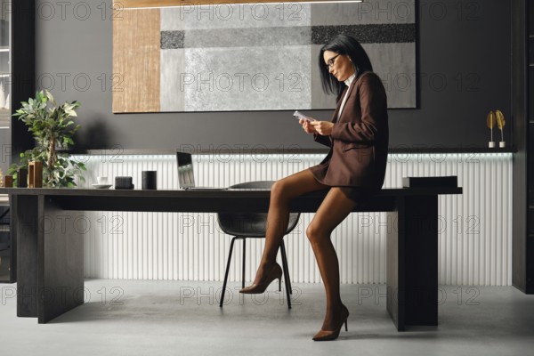 Serious business woman in a brown suit sits on a desk, focused on her smartphone. She checks her incoming messages