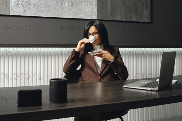 A real estate agent in a brown suit sits at a desk, sipping coffee from a small cup in a contemporary office. Her laptop is open, but she decided to make a coffe-break
