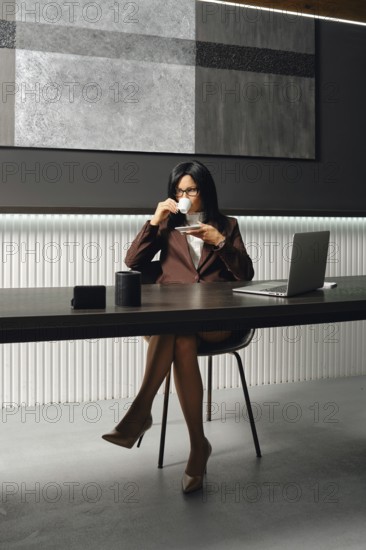 A confident businesswoman is seated at a sleek desk in a contemporary office. She sips coffee while working on a laptop, surrounded by minimalist decor, showcasing a productive atmosphere