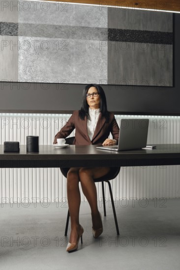 A slender woman with glasses and a stylish suit sits at an office desk with opened laptop. She stares sternly at the camera