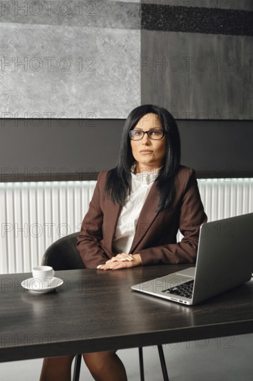 A focused businesswoman dressed in formal attire sits at a desk in a modern office environment. She appears to be engaged in a thoughtful discussion, with a laptop and coffee cup in front of her