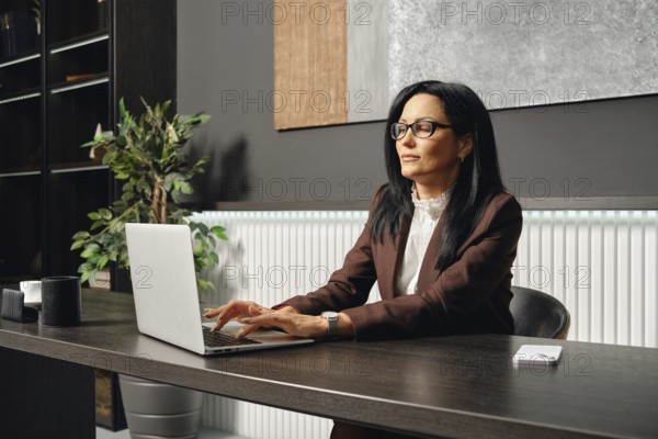 A businesswoman sits at a desk and typing on her laptop in an office. The space features elegant decor, including a plant and a minimalist color palette, enhancing productivity