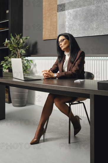 A woman dressed in business attire sits at an office desk in office and looking at the screen of laptop during a video call