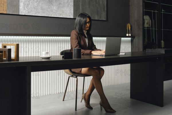 A businesswoman sits at a desk in a stylish office, focused on her laptop screen