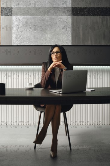 A businesswoman sits at a sleek desk in a contemporary office, dressed in strict suit. She gazes thoughtfully above her laptop and thinking on new strategy