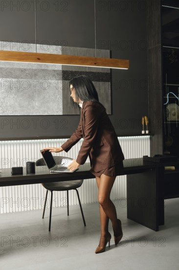 A confident professional woman in formal attire arranges her laptop on a sleek desk in a stylish office. The space features contemporary decor with a minimalist design and elegant lighting