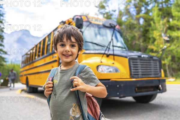 Happy schoolboy carrying backpack is waiting for the school bus in front of blurred mountain landscape, ready to start a new educational year