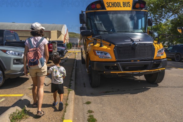 Mother and son walking hand in hand towards a bright yellow school bus in a bustling parking lot, embracing the excitement of a new school year on a warm, sunny day