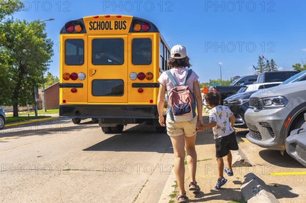 Mother walking her son to the school bus on the first day back to school, holding hands and heading towards the yellow bus parked near the school