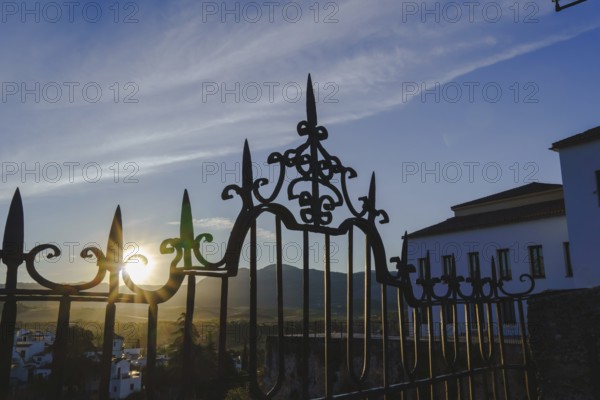 Sun shining through the ornate wrought iron gate at sunrise in ronda, spain, with mountains and white houses in the background in ronda, malaga, spain
