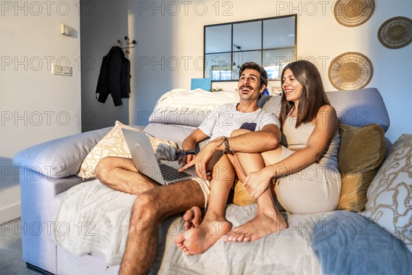 Young couple laughing while watching a streaming movie on their laptop, sitting comfortably on the sofa in their modern apartment, enjoying a relaxing evening together
