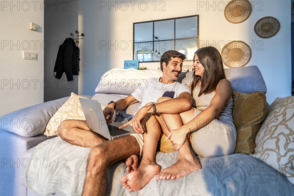 Young couple relaxing on the sofa at home, browsing on a laptop while enjoying the warm glow of sunset light streaming through the window