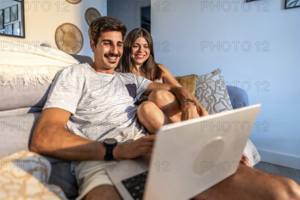 Young couple relaxing on sofa, enjoying streaming movie on laptop in cozy living room, illuminated by warm sunlight
