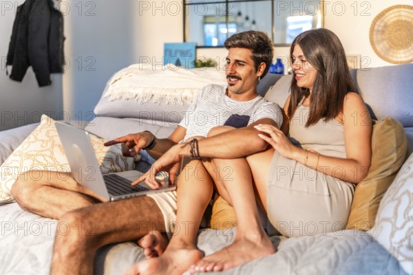Young couple relaxing on comfortable sofa, browsing internet on laptop, enjoying time together in cozy living room
