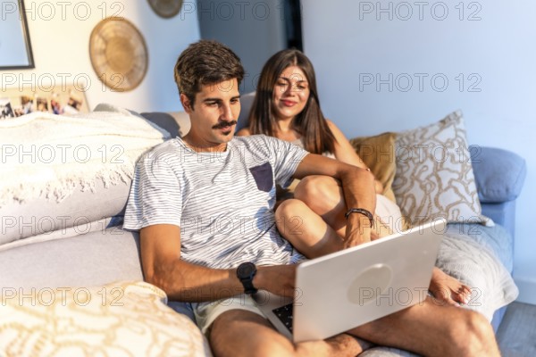 Young couple sitting comfortably on sofa browsing internet on laptop, enjoying time together at home