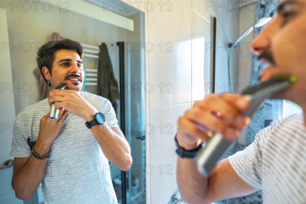 Young man trimming his beard using an electric razor, looking at himself in the bathroom mirror during his morning routine