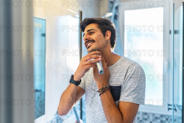 Happy young man trimming his beard in the bathroom mirror using an electric razor, enjoying his morning routine