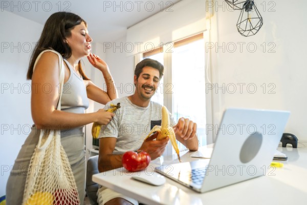 Young couple smiling and enjoying bananas while working on a laptop in a bright, modern home office, embracing a healthy lifestyle together