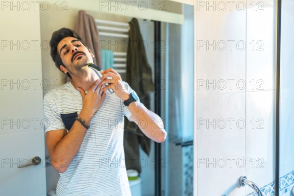 Young man carefully shaving his neck with an electric razor, looking in the bathroom mirror during his morning routine