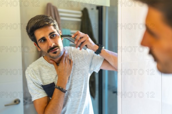 Concentrated young man trimming his beard with electric razor in bathroom mirror, morning routine, personal hygiene and care concept