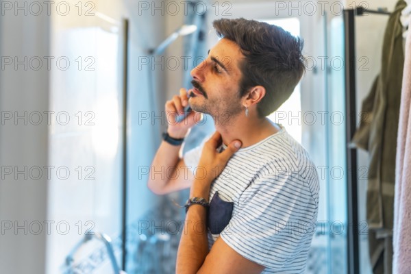 Young man trimming his mustache with an electric razor and touching his neck while looking in the mirror in a modern bathroom