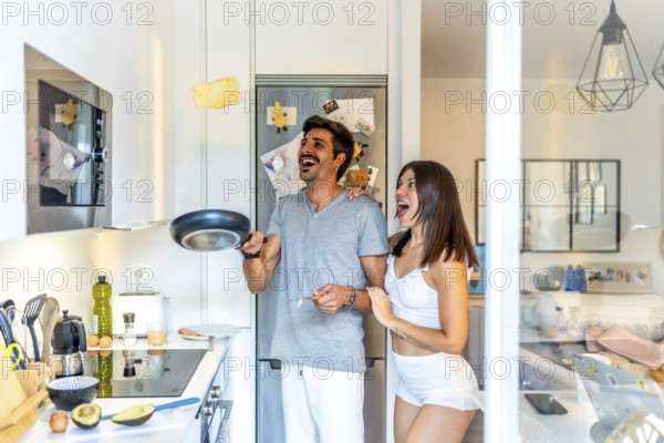 Cheerful young couple having fun preparing breakfast, flipping omelette or pancake in the air in a modern kitchen